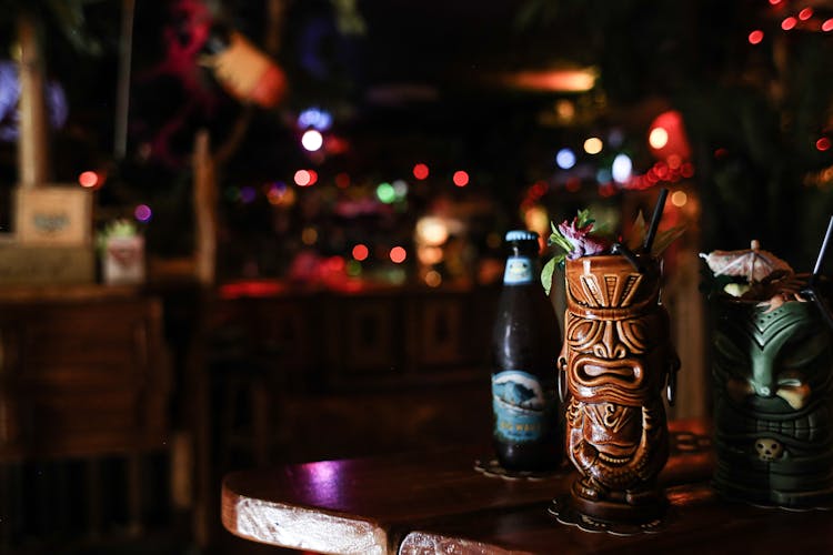 Beer Bottle And Ceramic Tiki Mugs On Wooden Table