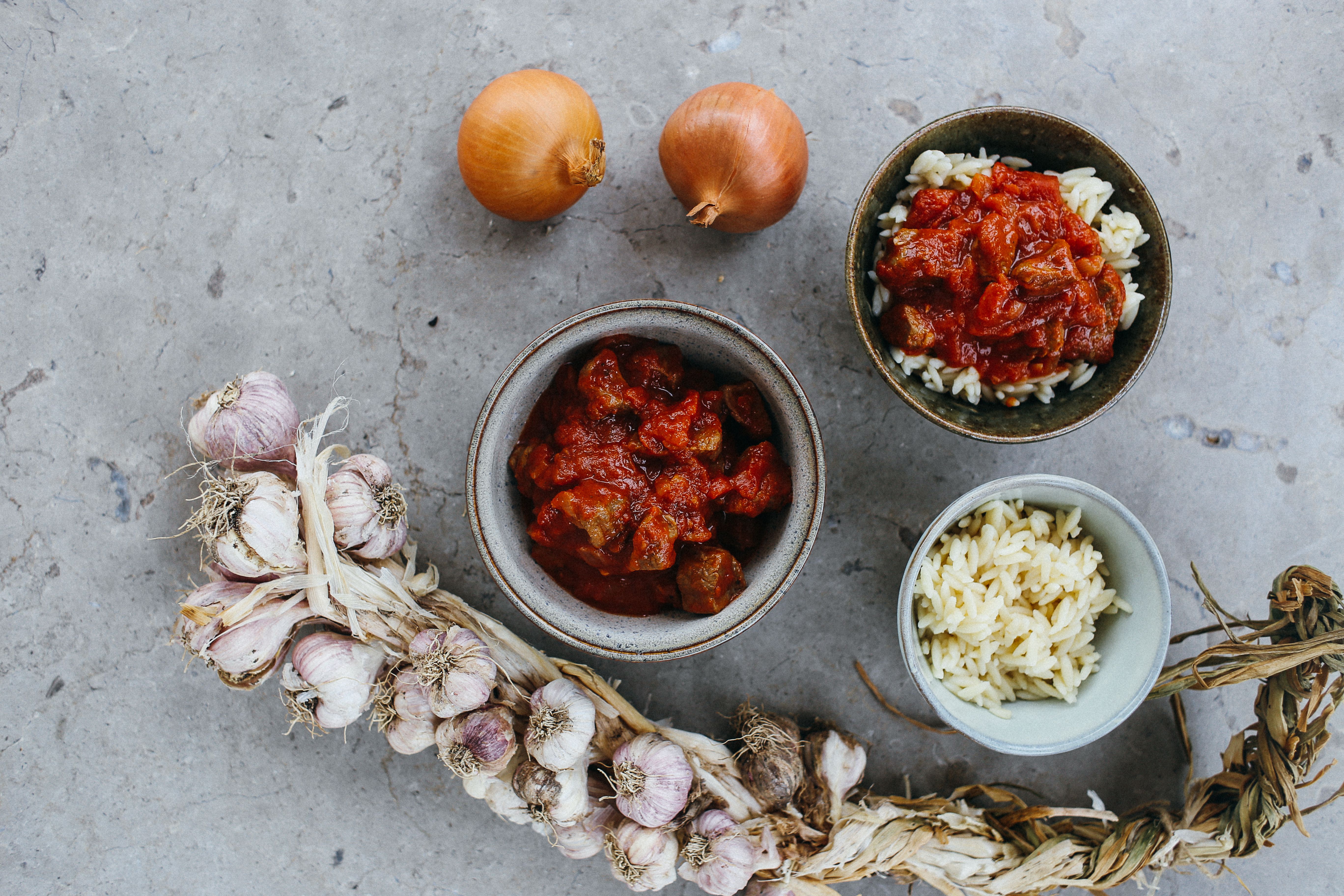 Top view of a rustic meal with garlic, onions, and bowls of rice and stew on a stone surface.