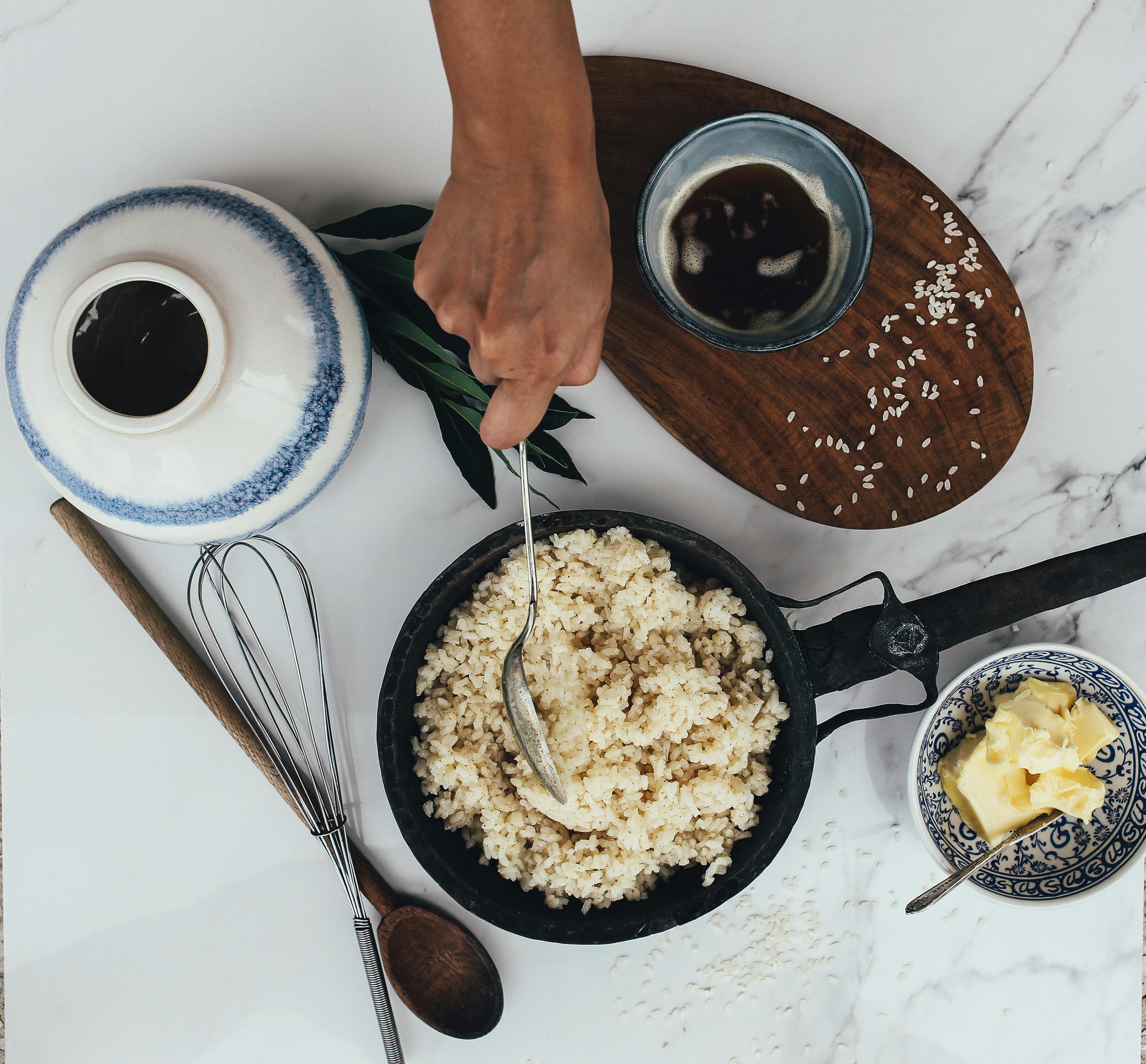 Hand stirring rice in a pan with butter and tea on a marble surface for a homely meal setup.