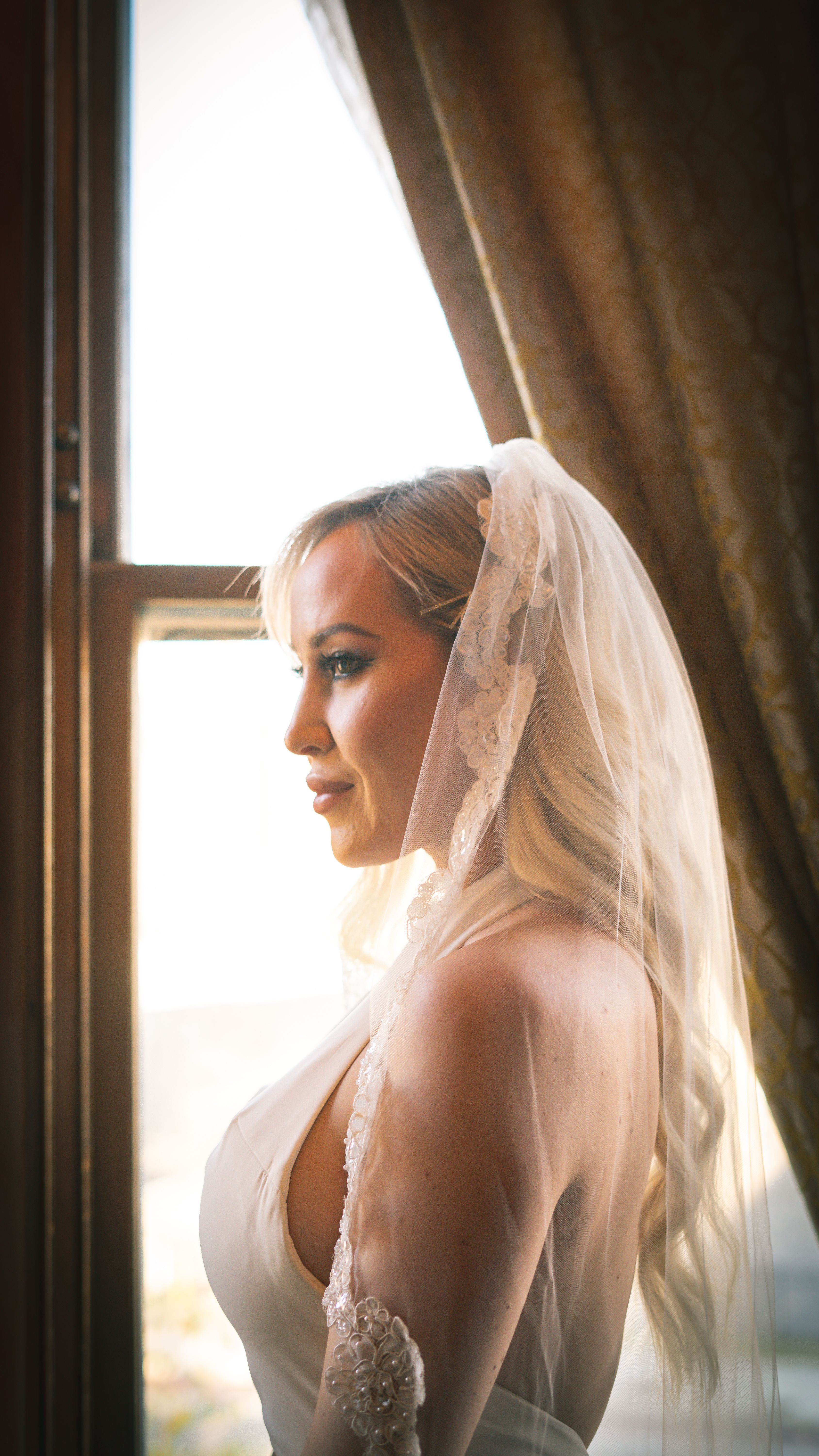 Side view of female in white wedding dress looking away near window decorated with brown curtain in sunshine
