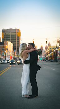 Side view of newlywed couple in elegant suit and white wedding dress with veil hugging on urban road