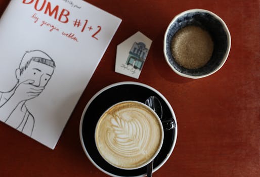 A cappuccino with latte art, a comic book, and a bowl of sugar on a red table.