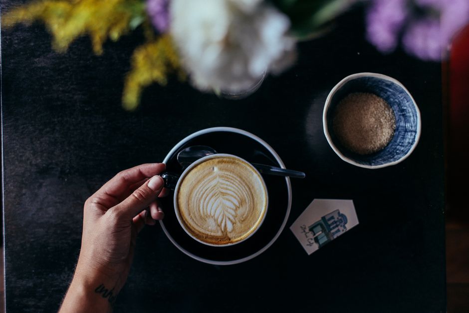 Hand holding a latte with art, accompanied by brown sugar in a bowl, on a dark table.
