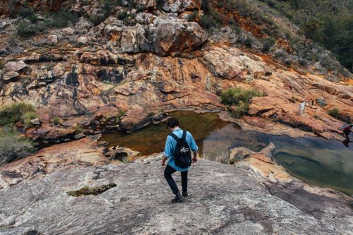 Free A man wearing a backpack explores rugged rocks beside a tranquil pond in a mountain landscape. Stock Photo
