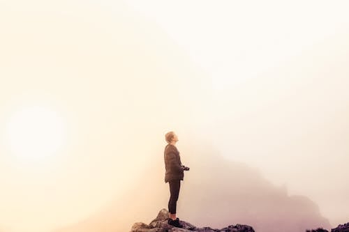 Man in Black Jacket Standing on Rock Formation Holding a Camera on a Foggy Day