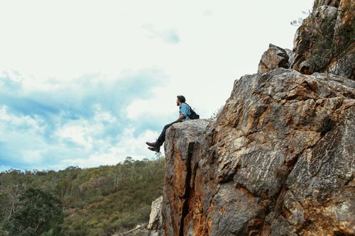 Free A man sits on a rocky cliff, admiring the scenic valley beneath a dramatic sky. Stock Photo