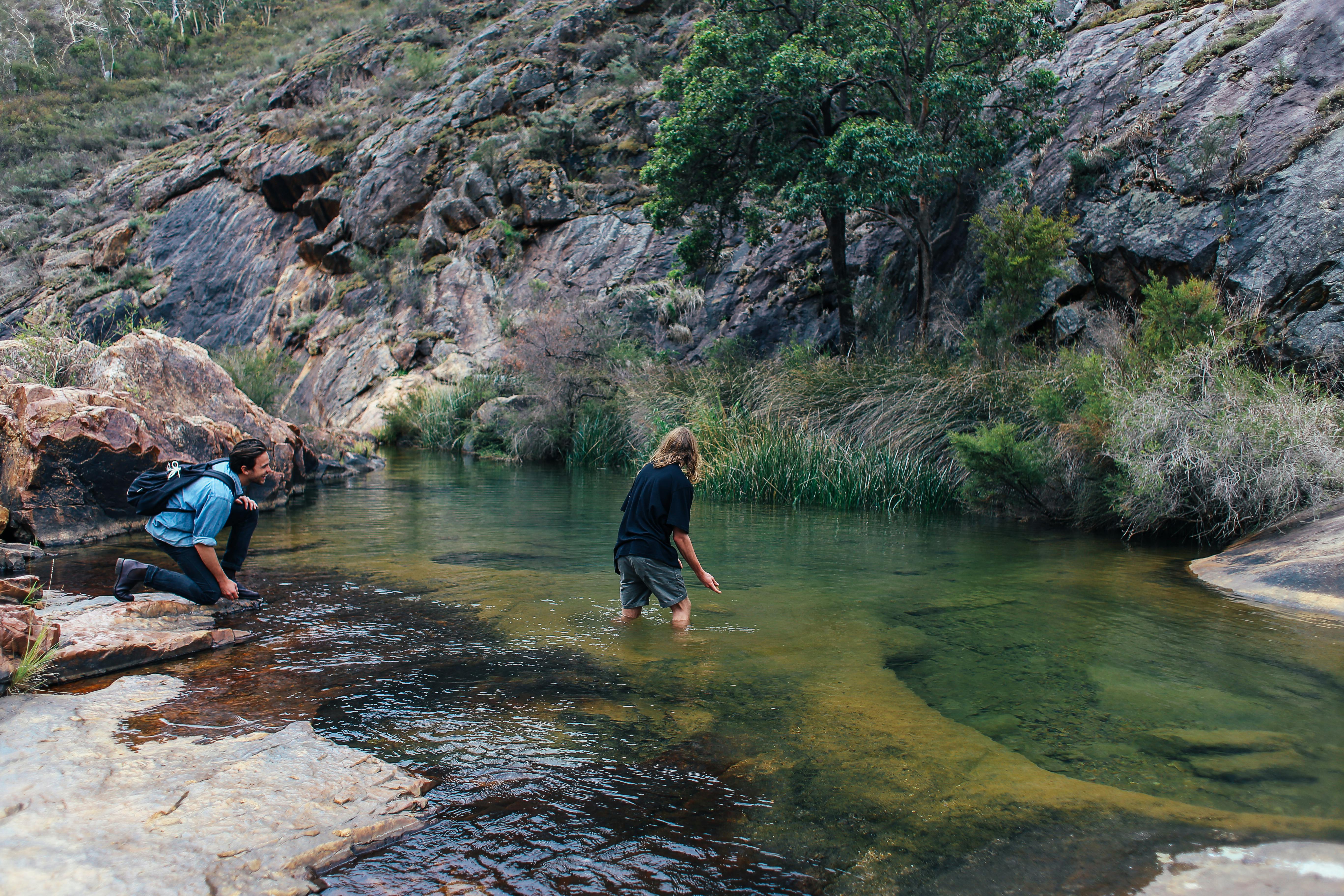 Two hikers explore a shallow creek surrounded by rocky terrain and lush greenery.