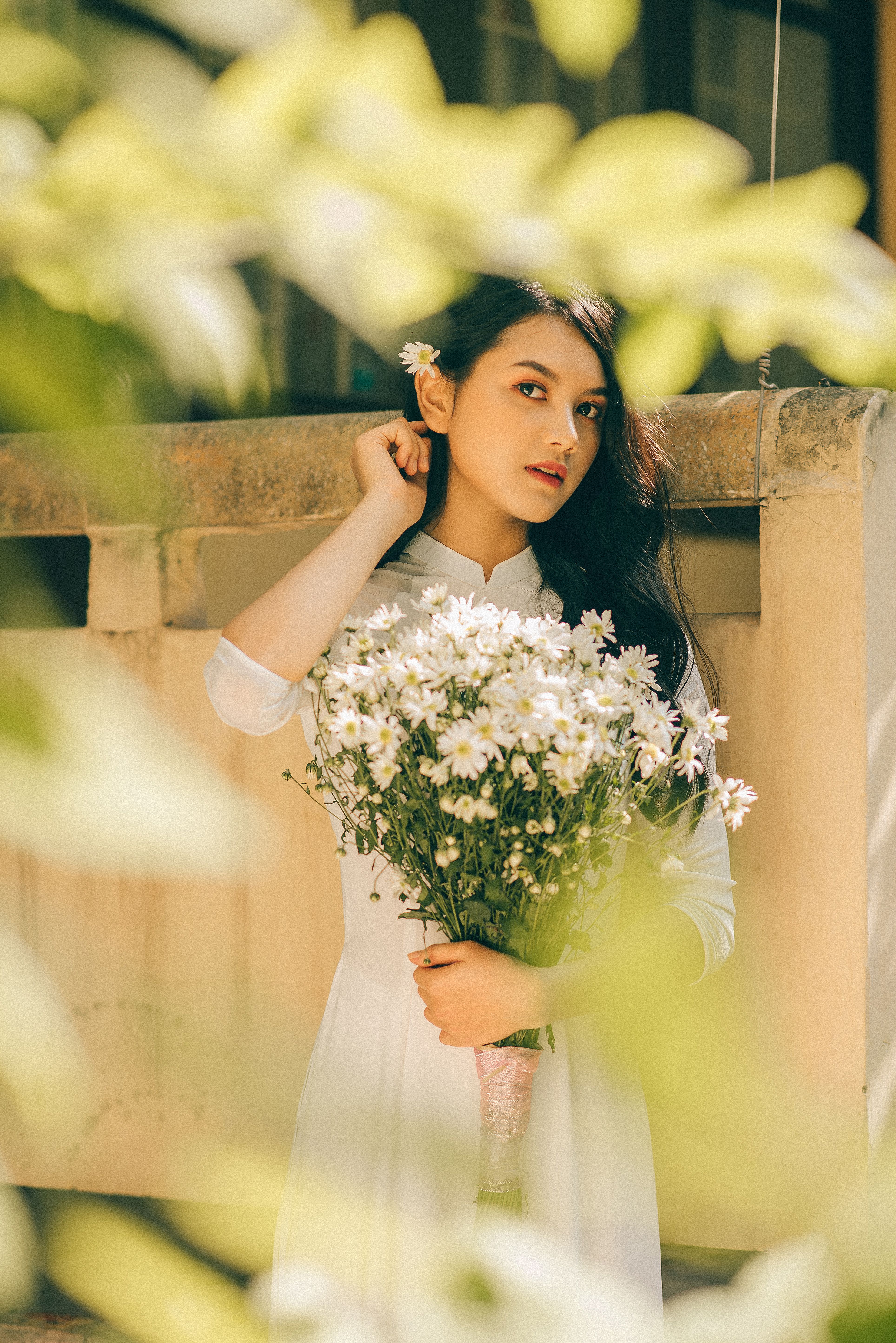 一位优雅的女人留着长发，在户外被绿叶环绕，手里拿着一束白雏菊
