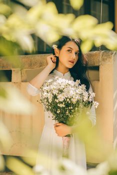一位优雅的女人留着长发，在户外被绿叶环绕，手里拿着一束白雏菊