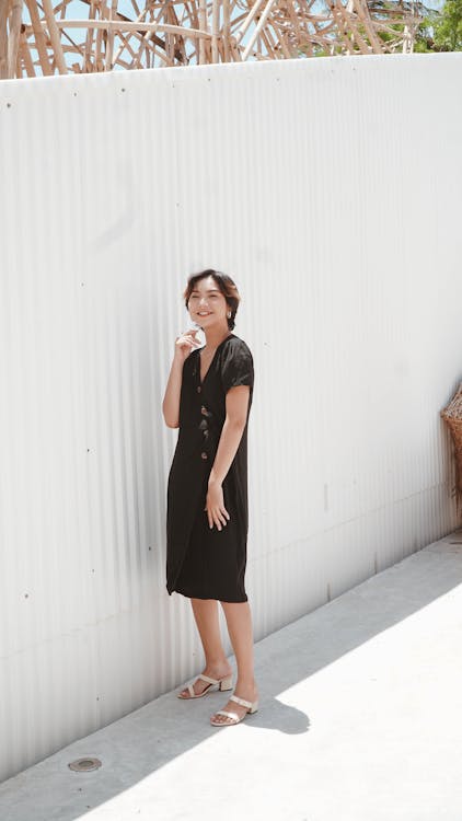 Free Woman in Black Dress Standing on Concrete Pavement Near White Wall Stock Photo