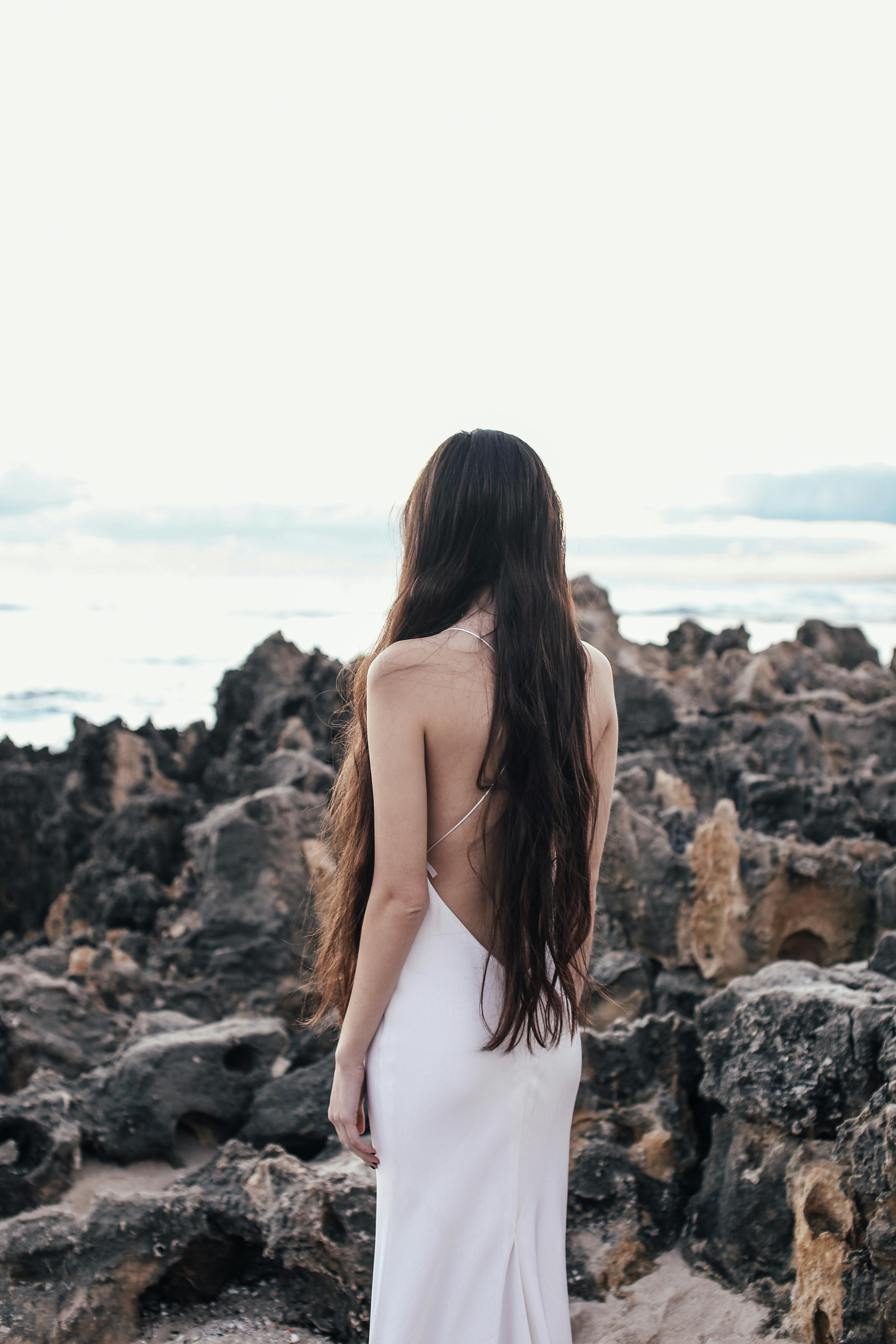 一个长头发的女人，穿着白色连衣裙，站在岩石岸边，面朝大海