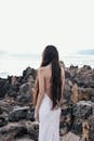 一个长头发的女人，穿着白色连衣裙，站在岩石岸边，面朝大海