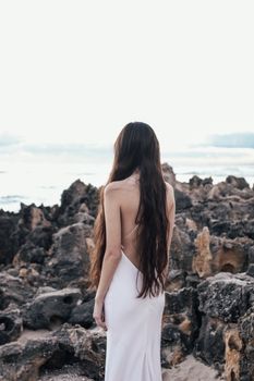 一名长发女子身着白色连衣裙，站在岩石海岸边，面朝大海