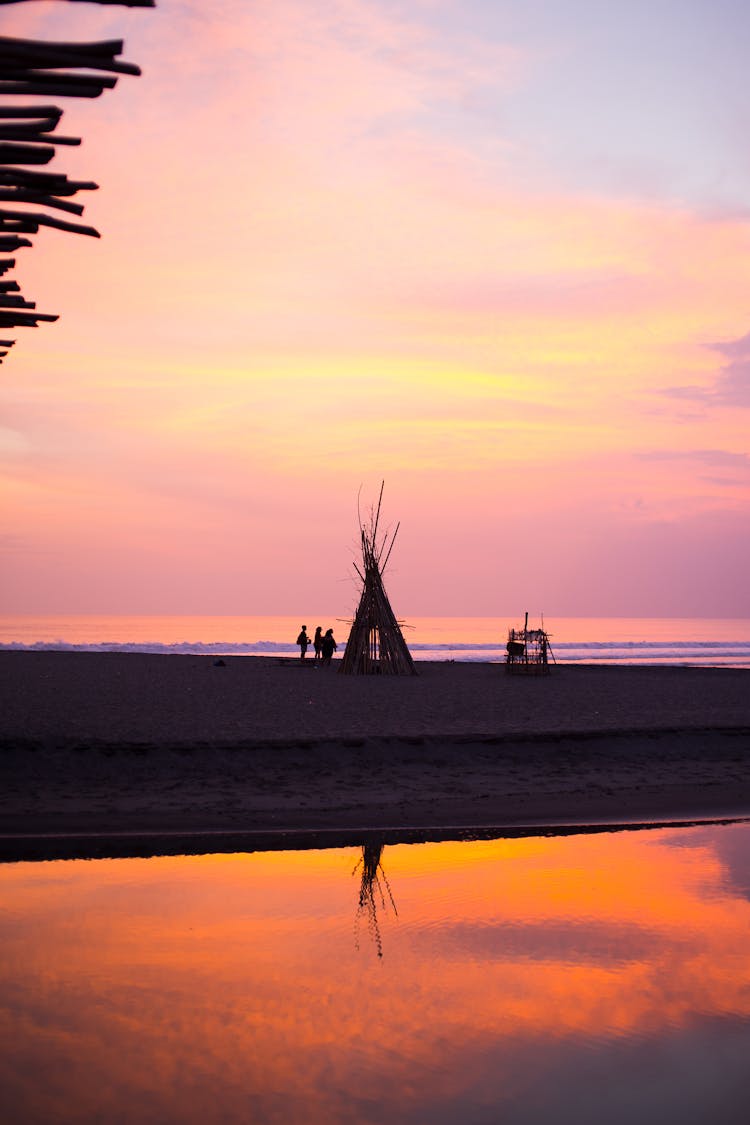 Silhouette Of People Standing Near A Teepee On Beach During Sunset