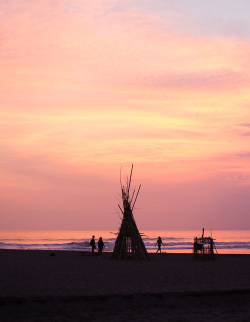 Free Silhouetted scene of people near beach teepee structures under a vibrant sunset sky. Stock Photo