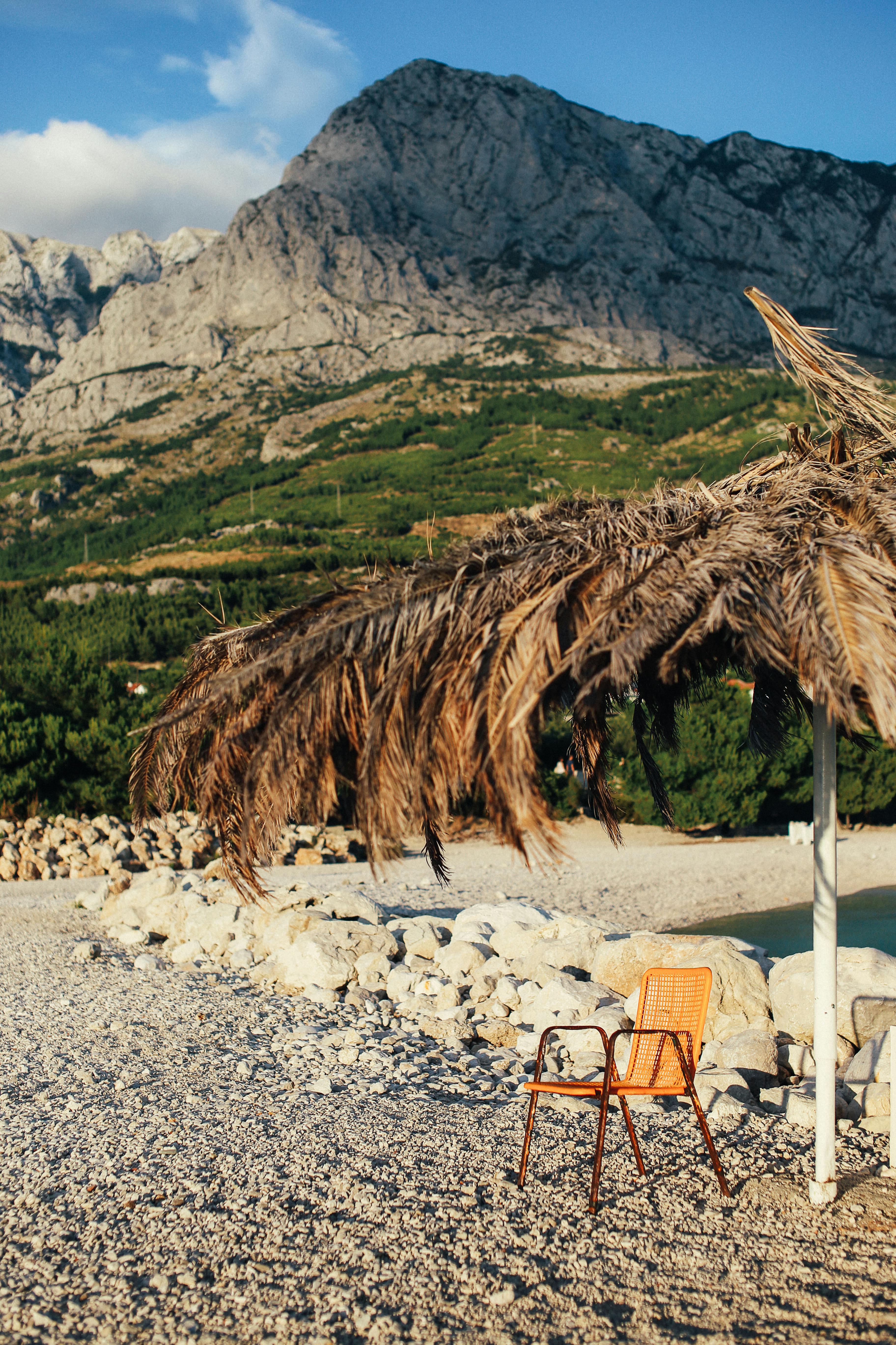 宁静的海滩上，棕榈叶的阴凉里放着一把空椅子，背靠着山脉