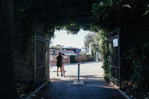 Free A person with a bicycle standing at a leafy city gate leading to a sunny street. Stock Photo
