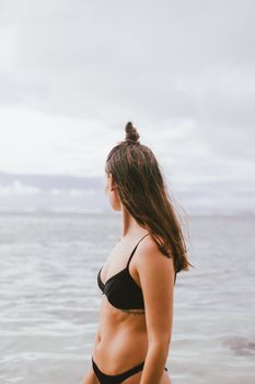 A woman in a black bikini stands by the ocean, gazing at the horizon under a cloudy sky.