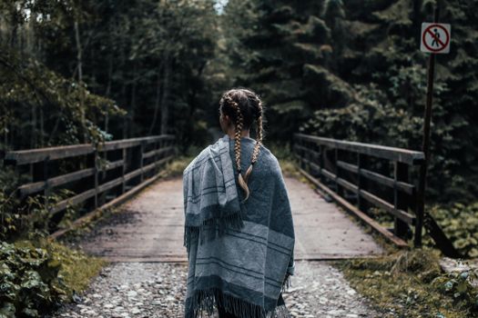 一名扎着辫子的妇女裹着披肩，站在森林中郁郁葱葱的桥上