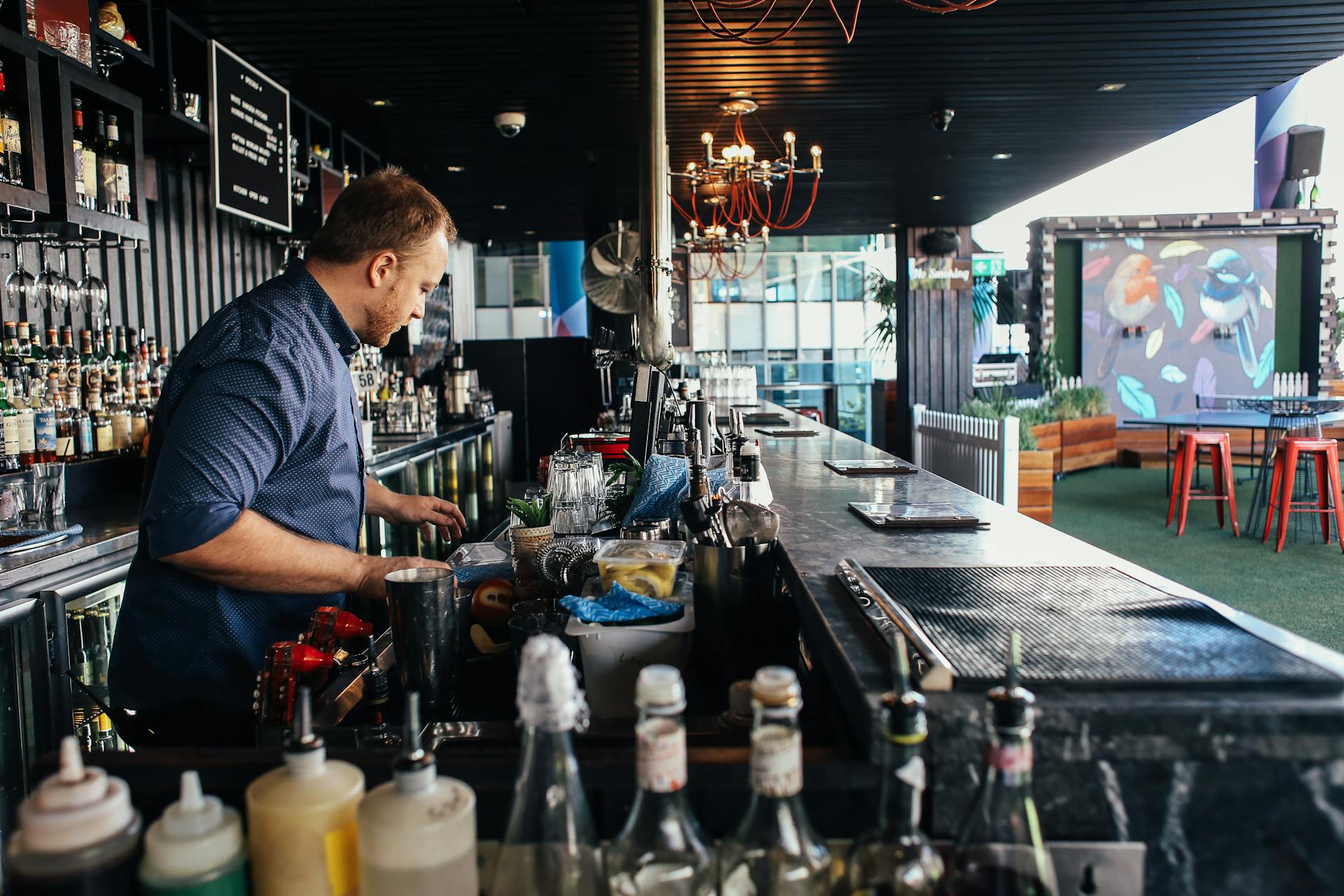 A bartender mixes cocktails at an outdoor rooftop bar with bottles on display.