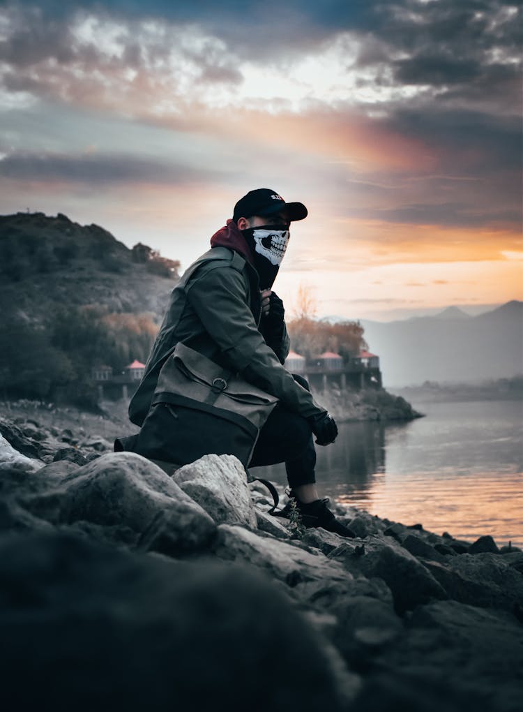 Man In A Skeleton Face Mask And Black Cap Sitting On A Rocky Coast At Dusk 