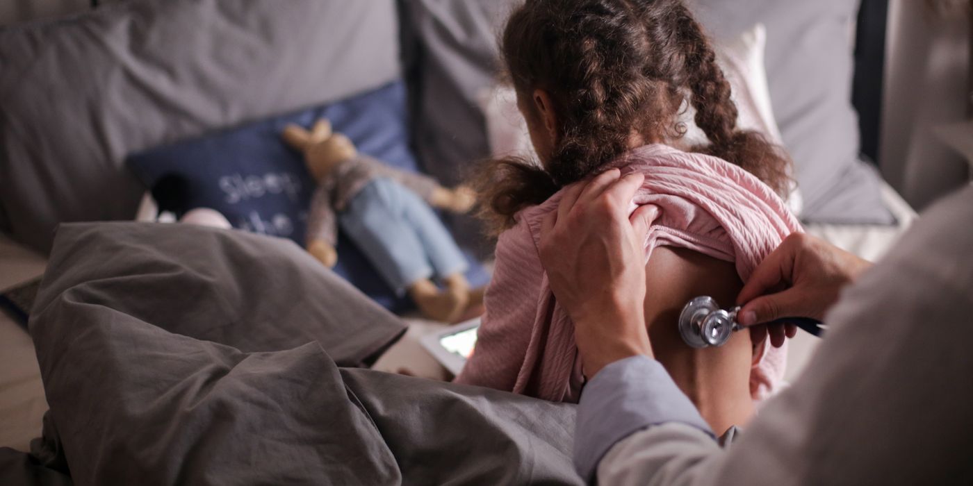 A pediatric doctor checks a child's back with a stethoscope in a cozy bedroom setting.