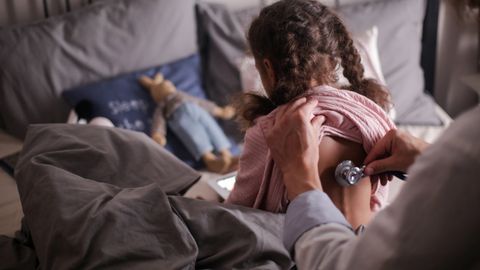 A pediatric doctor checks a child's back with a stethoscope in a cozy bedroom setting.