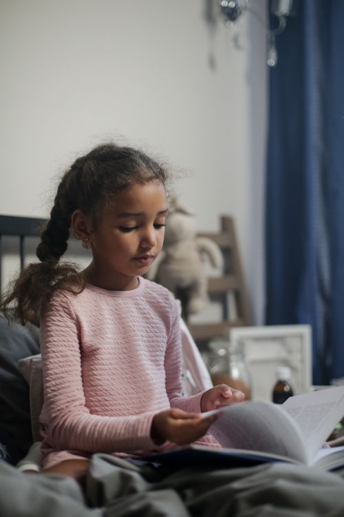 Free A young girl in a pink dress sits on a bed reading a book, creating a cozy indoor scene. Stock Photo