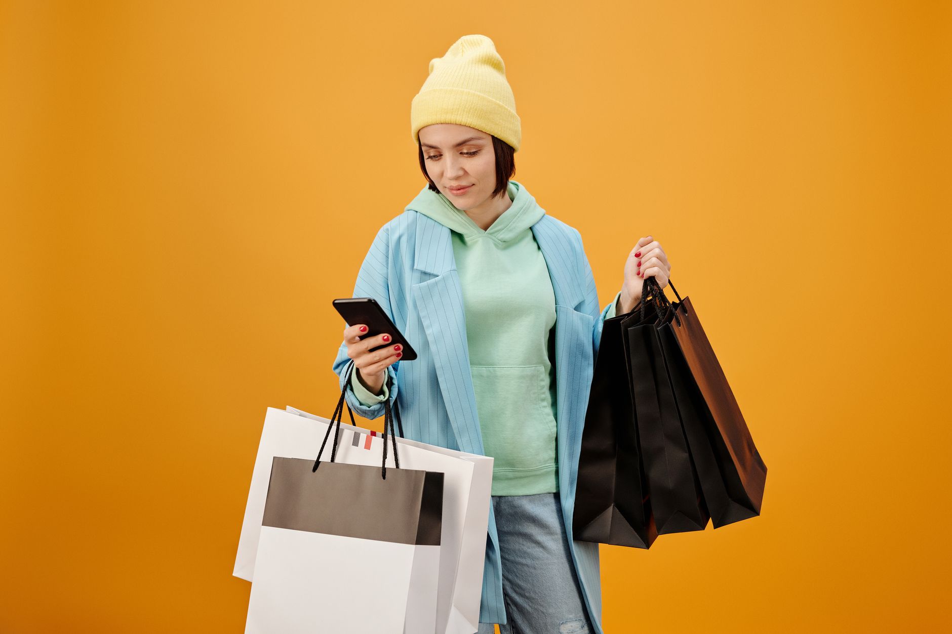 Stylish young woman with paper bags, checking her smartphone after shopping spree on colorful background.