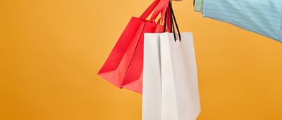 A hand holds several colorful paper shopping bags against a bright yellow background.