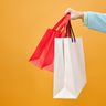 A hand holds several colorful paper shopping bags against a bright yellow background.
