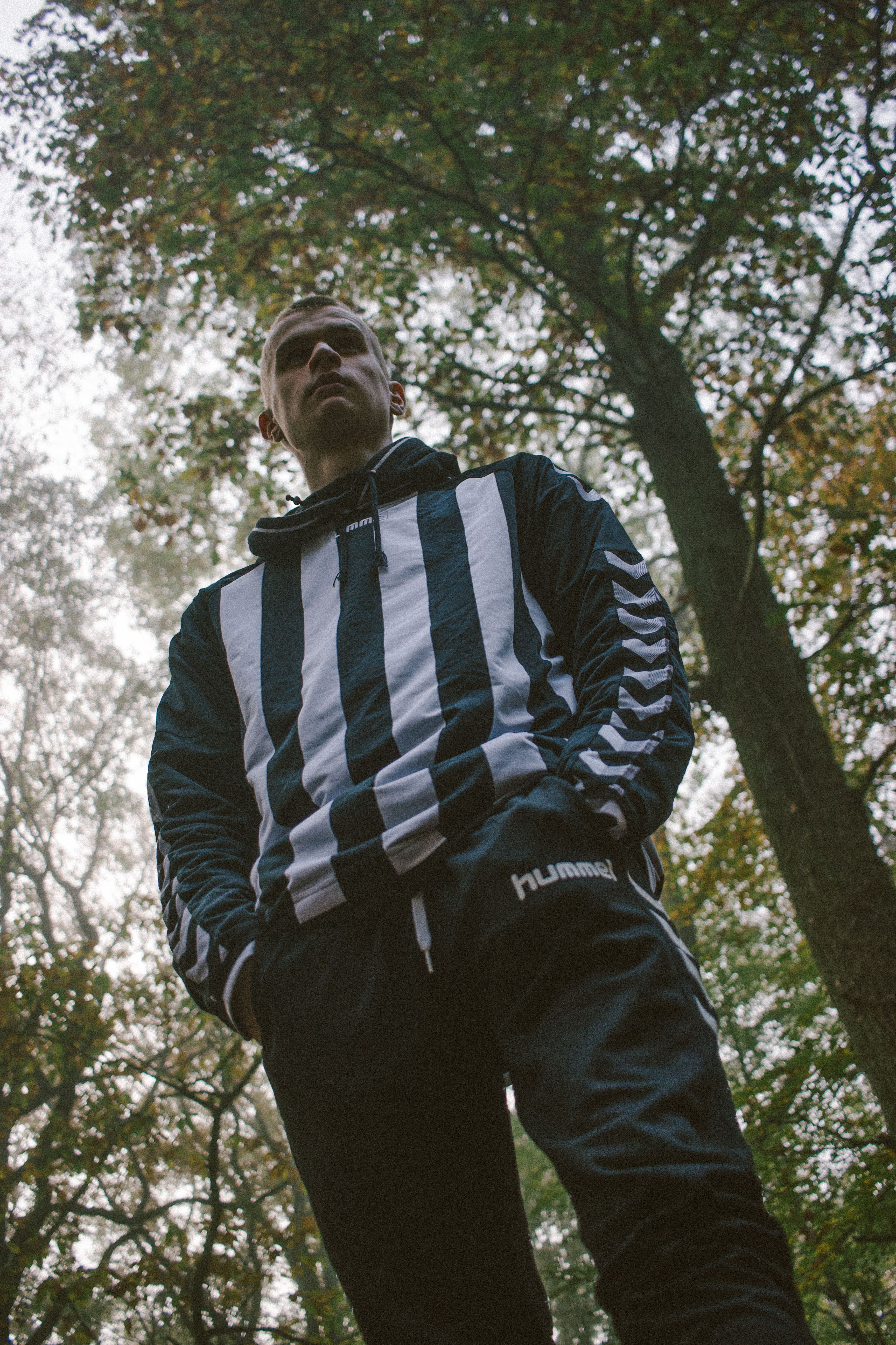 From below of young guy in ornamental hoodie with hands in pockets looking away near green trees