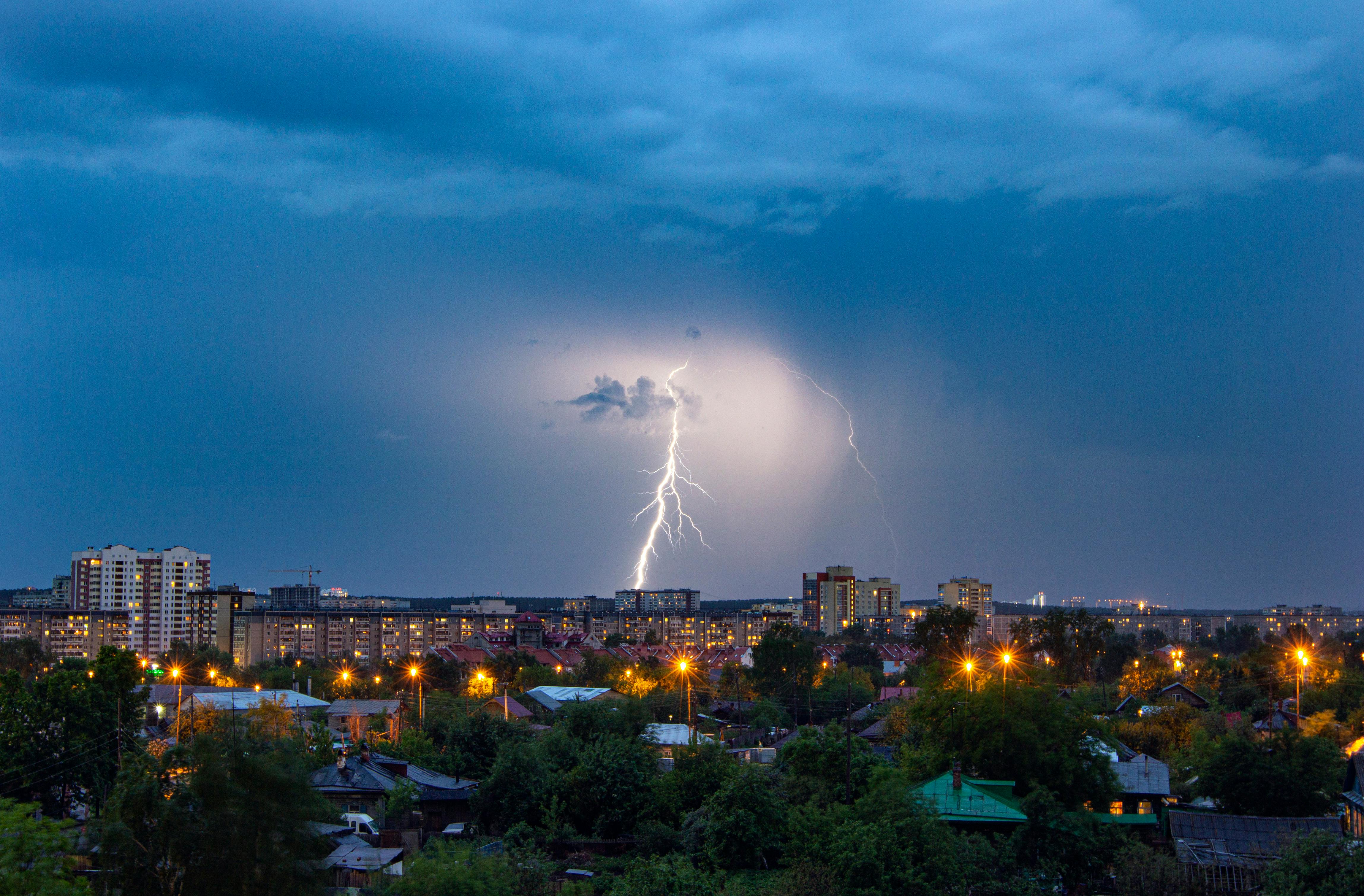 Bright lightning shining near town with houses near green trees and streetlamps under sky in evening