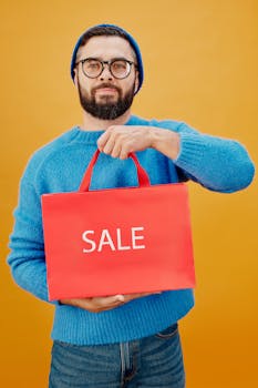 Man with beard and glasses in blue sweater holding red sale shopping bag against yellow background.