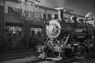 Black and white fragment of old steam locomotive with chimney on railroad near building and leafless tree in city on street