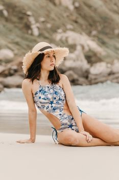 Young feminine tourist in trendy swimwear and hat resting on ocean beach against mountain and looking away