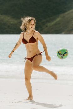 Young tattooed female tourist in swimwear with raised leg playing with ball on sandy sea beach