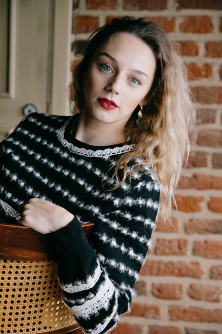 Young charming blond female with red lips in fluffy sweater resting on chair while looking at camera near brick wall