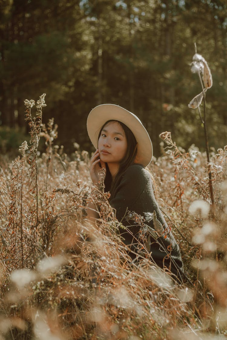 Charming Asian Woman In Hat Resting On Grassy Meadow