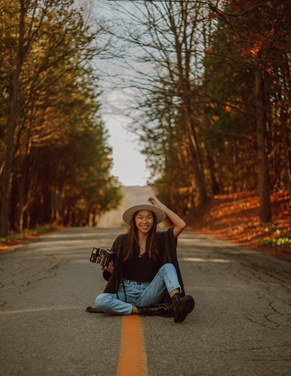 Full length delighted young Asian female wearing casual clothes and hat sitting on asphalt roadway with retro photo camera between yellow autumn trees