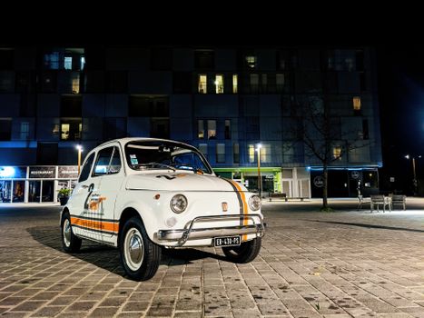 A classic Fiat 500 parked in a modern urban area at night, illuminated by street lights.