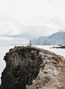Distant unrecognizable female traveler relaxing on rough rocky cliff and admiring spectacular sea view against cloudy sky