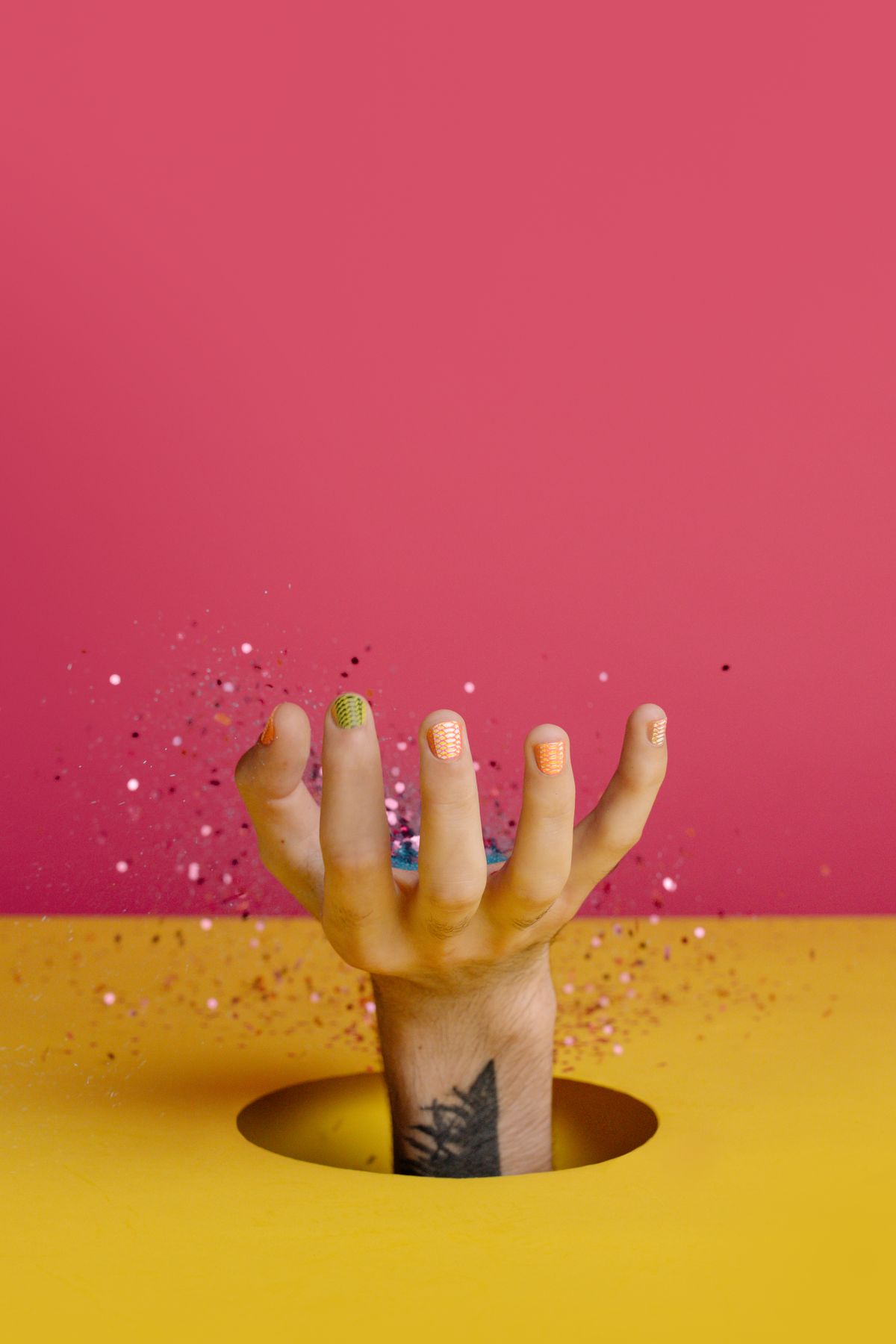 A hand with multicolored nails and tattoos reaching out of a hole with glitter against a pink background.