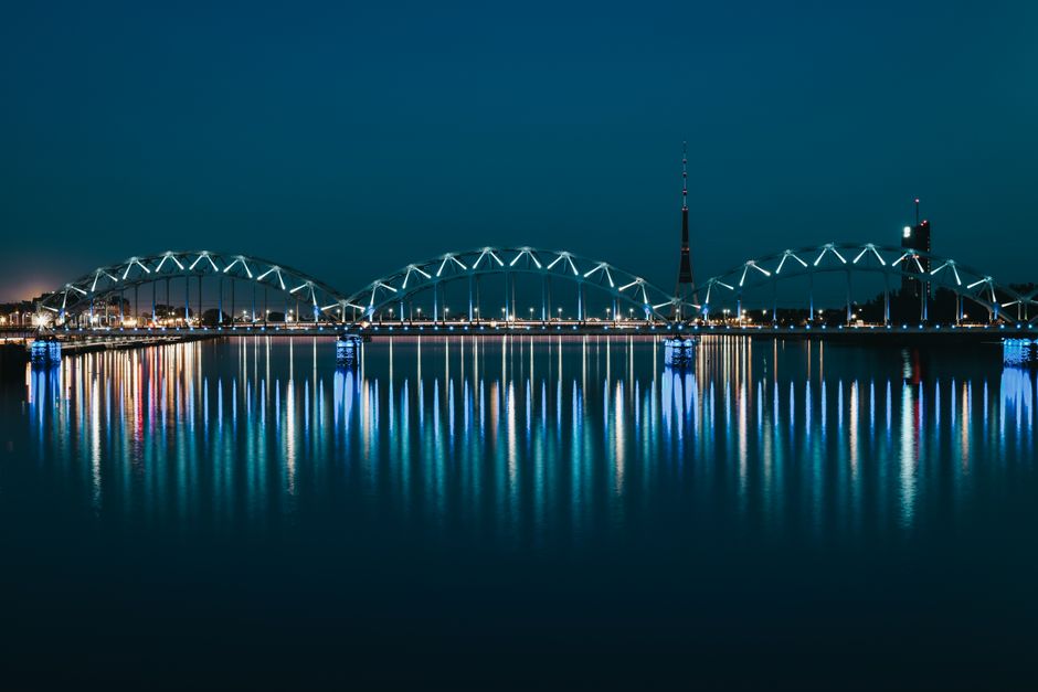 Illuminated historic arched iron Railway Bridge reflecting in river water against cloudless night sky in Riga