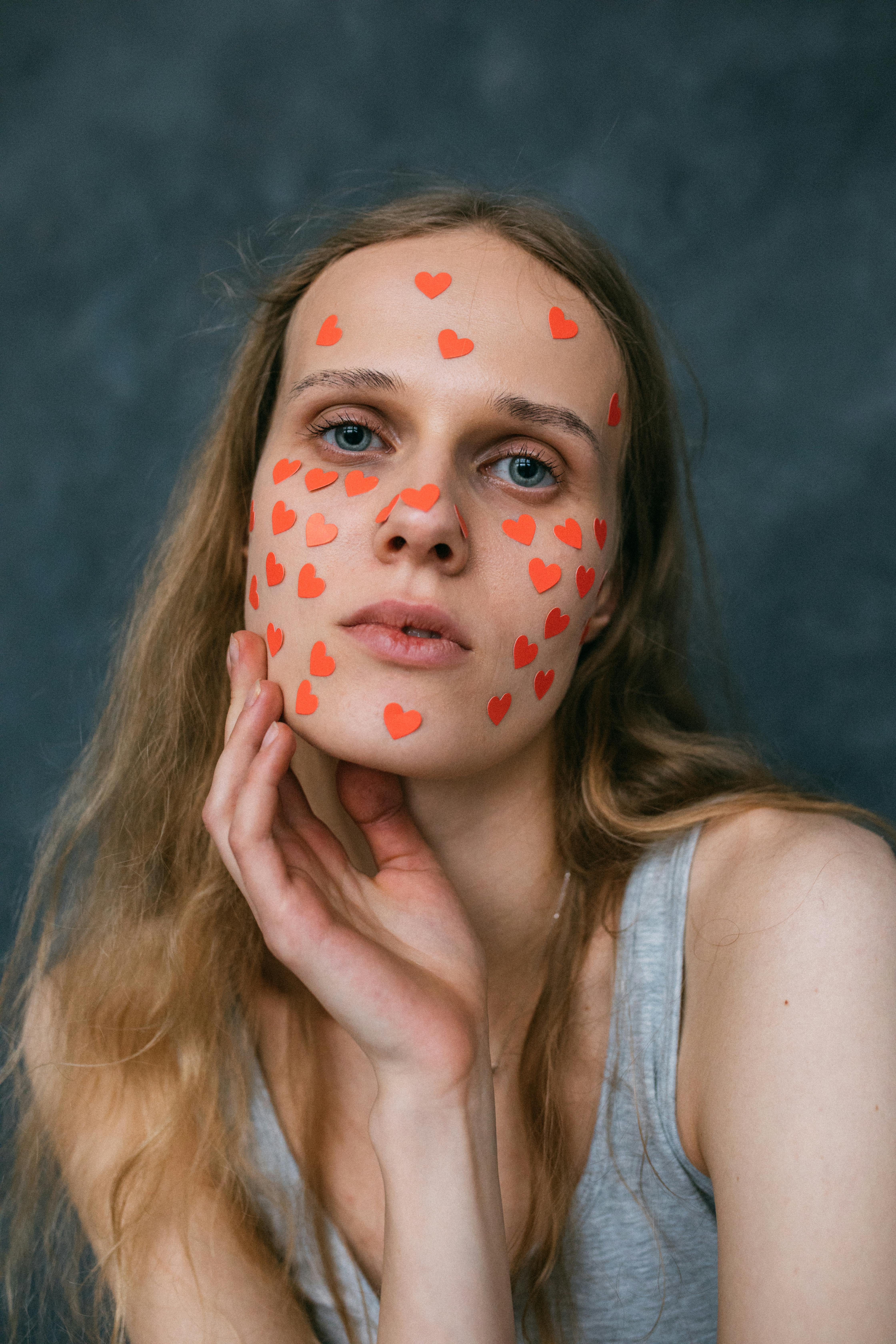 Close-up portrait of a woman with red heart stickers on her face, expressing a unique and artistic style.