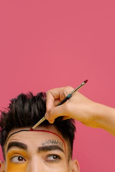 Close-up of creative makeup application on a man's forehead, featuring a pink background.