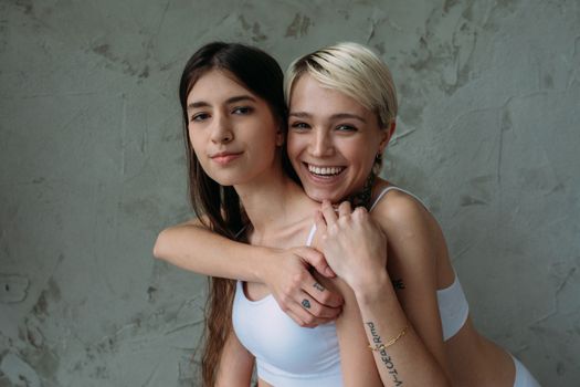 Two young women smiling and hugging against a textured concrete wall, expressing friendship and joy.