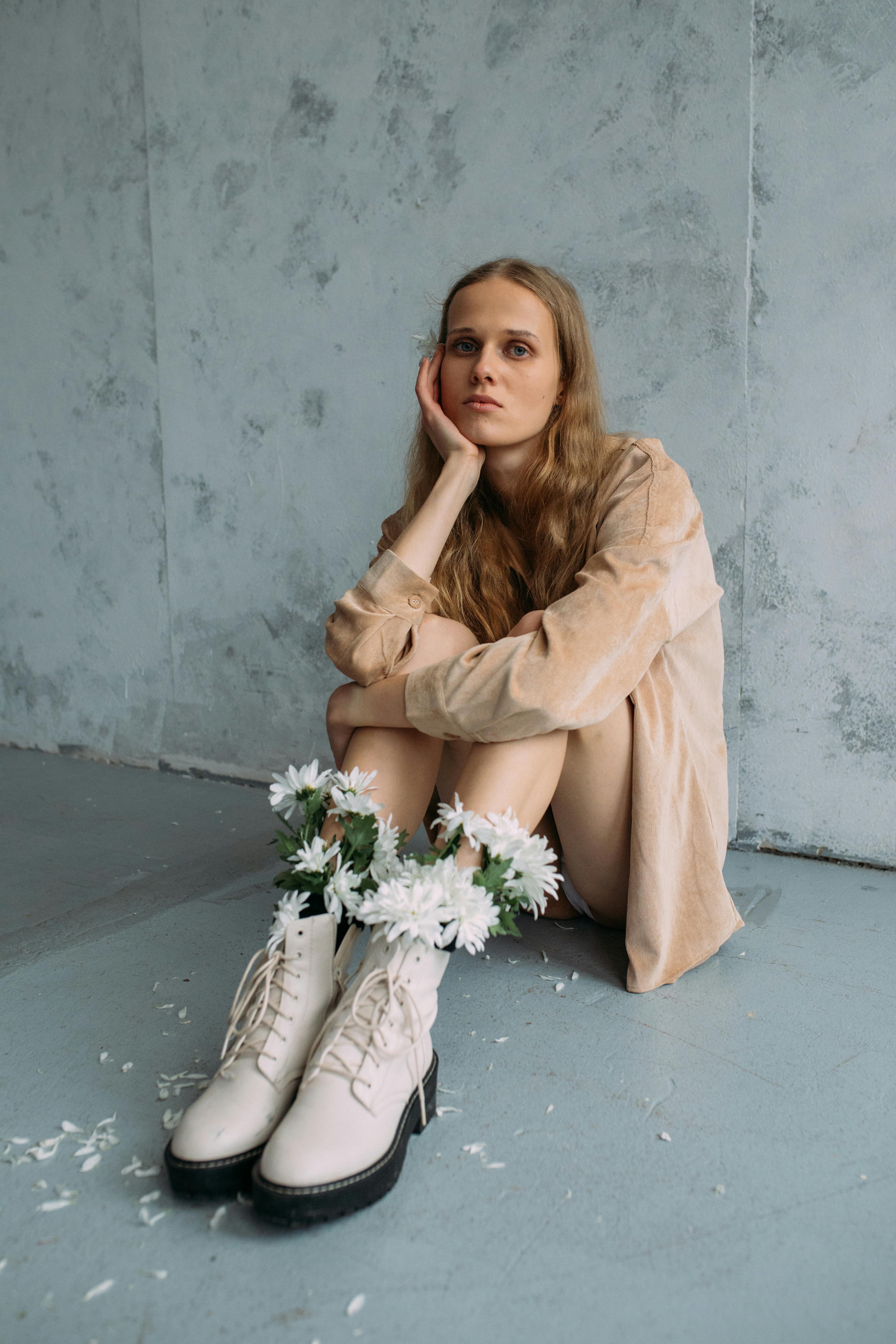 Woman sitting indoors, wearing white boots decorated with flowers, reflecting in thought.