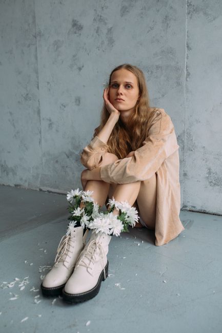 Woman sitting indoors, wearing white boots decorated with flowers, reflecting in thought.