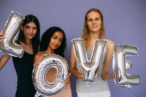 Free Three diverse women posing indoors with silver love letter balloons against a purple background. Stock Photo